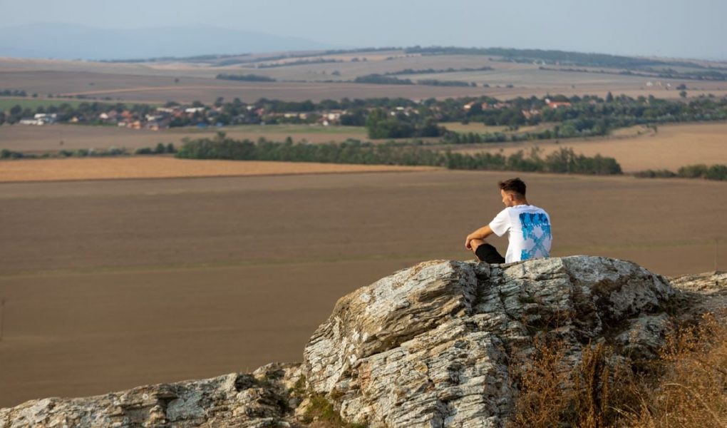 A man sits on a rock overlooking the vast countryside in a serene outdoor setting.
