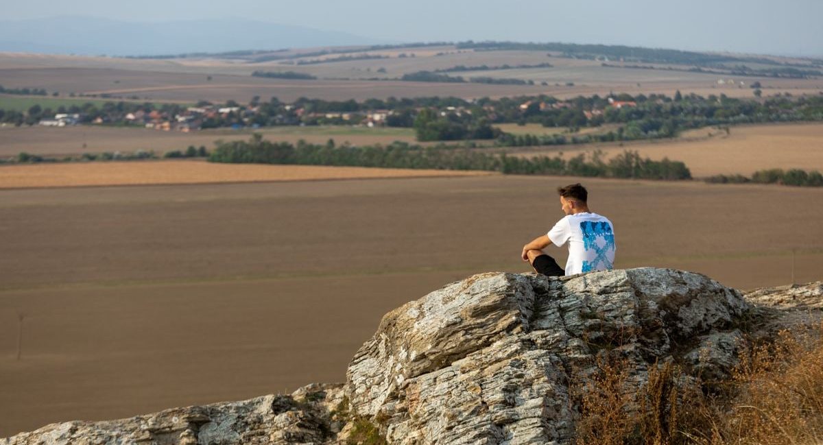 A man sits on a rock overlooking the vast countryside in a serene outdoor setting.